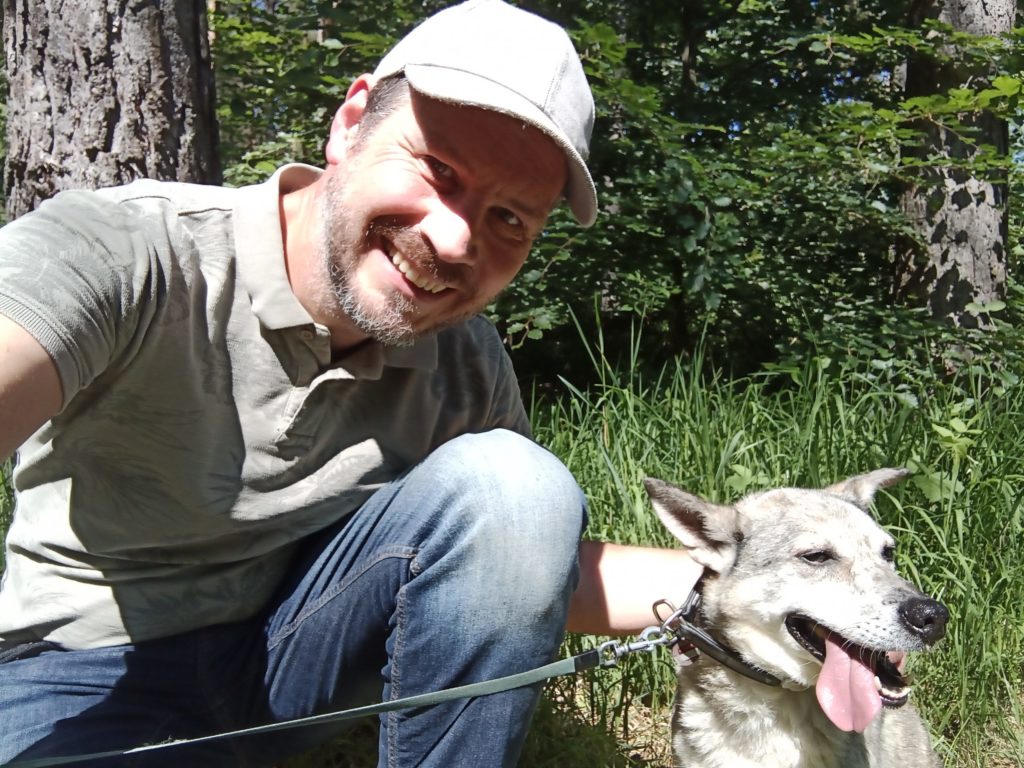 Jan Soeren kniet mit grauer Basecap und seinem Hund vor einem Waldhintergrund.