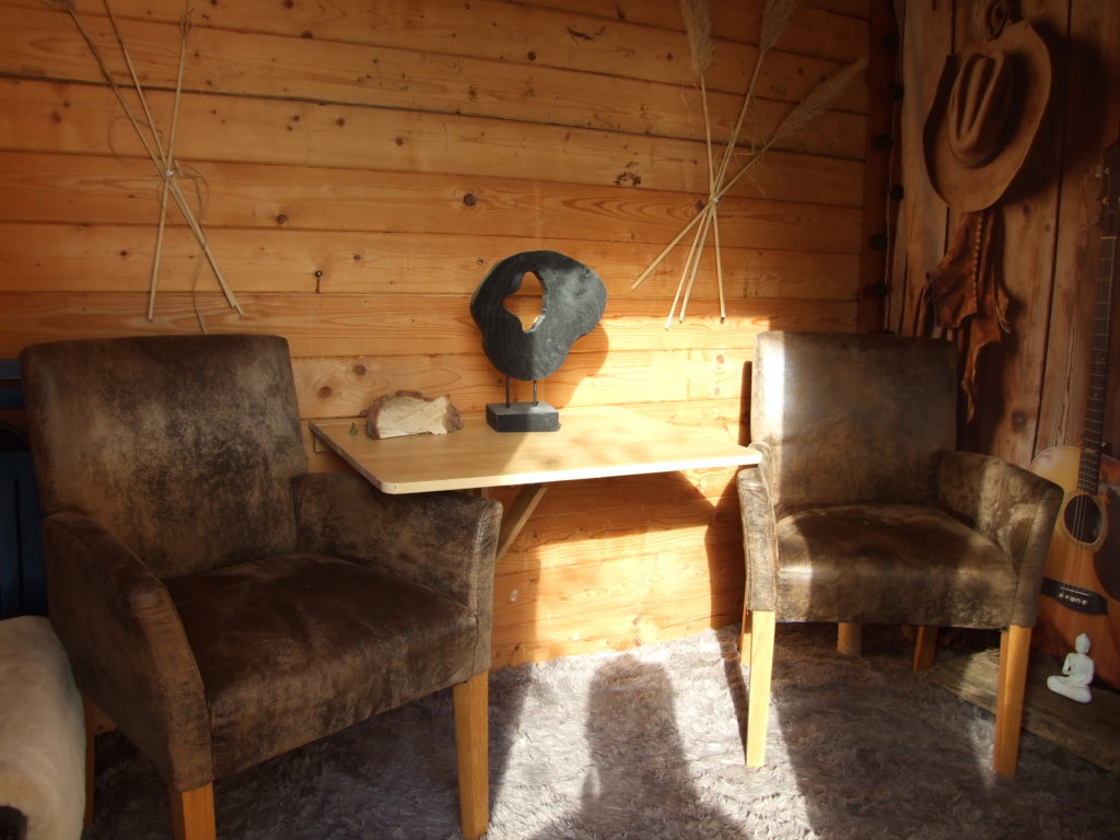 Beratungsraum. Gemütliches, rustikales Holzhaus mit zwei braunen Sesseln und einem Tisch mit schwarzer Holzskulptur.